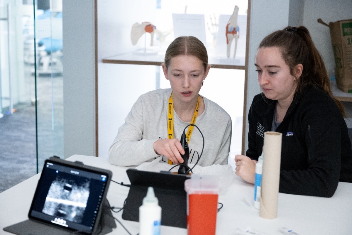 Medical student and high school student working with an ultrasound probe at a table.