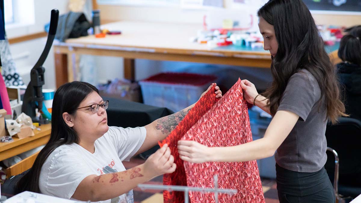 A woman standing holding a piece of fabric up to a woman seated holding a similar piece of fabric
