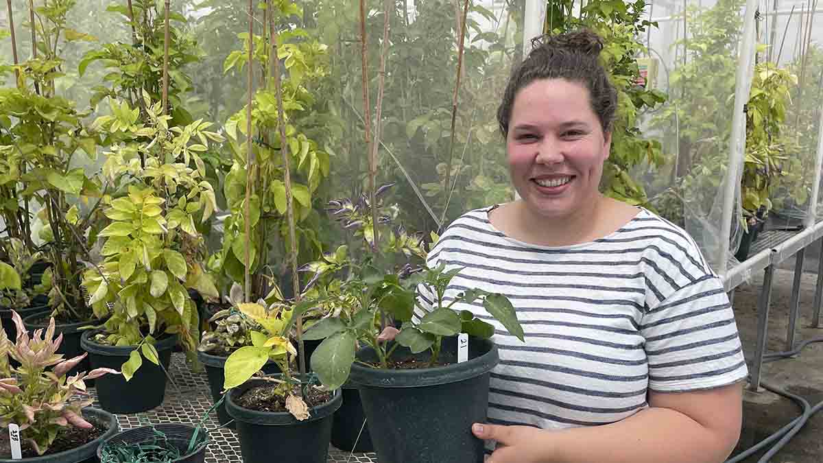 A woman holds a potted plant inside a greenhouse filled with plants.