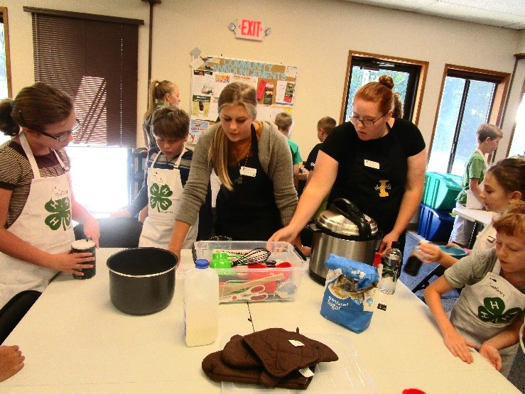 Youth of many ages wear aprons and gather around kitchen tools.