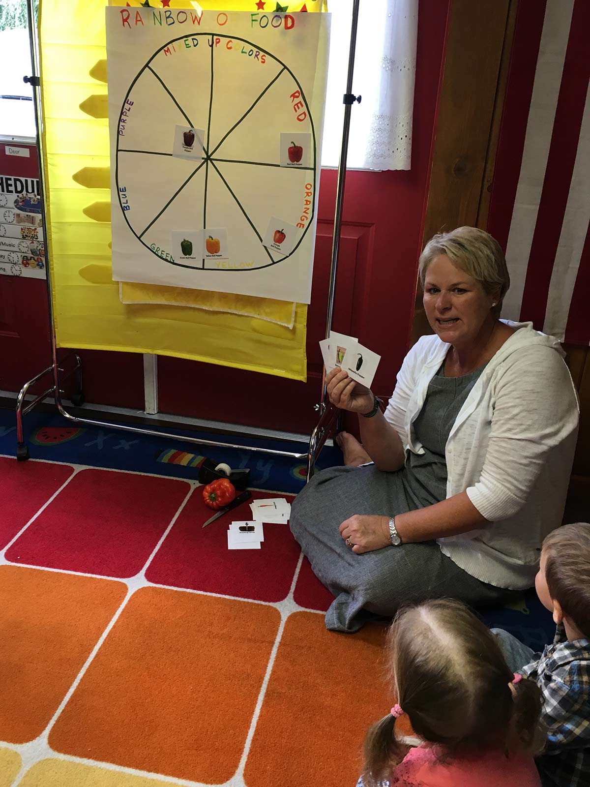 A woman holding up small cards in a classroom with a "rainbow of food" wheel.