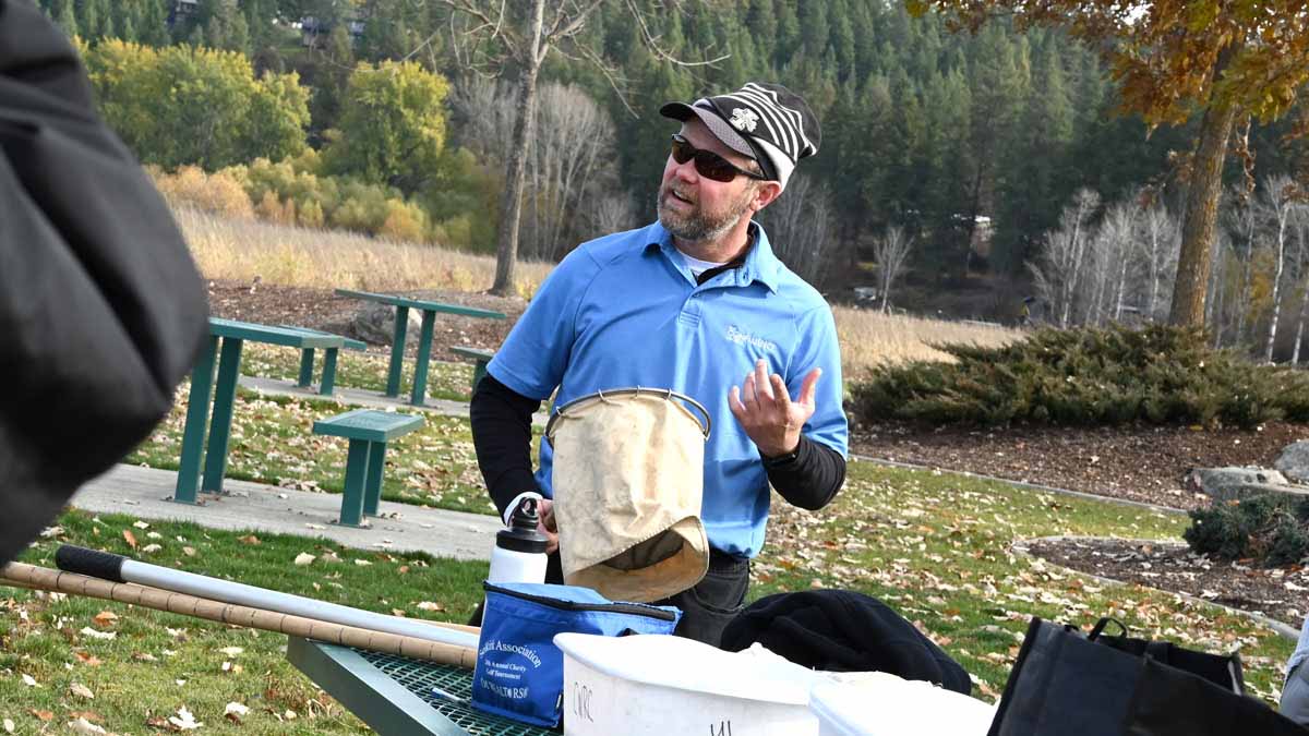 Jim Ekins stands at a table in a park holding a water net while talking to students out of the frame.