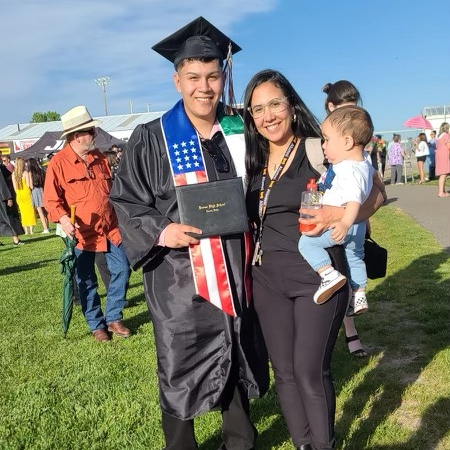 A man in a graduation cap and gown stands next to a woman holding an infant.