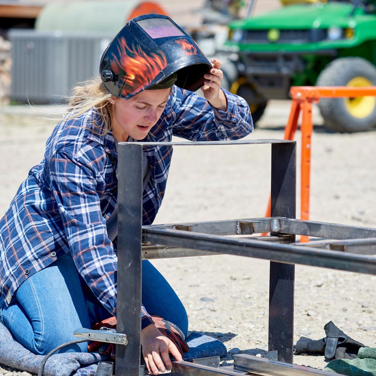 A student lifts her welding mask to inspect her work.