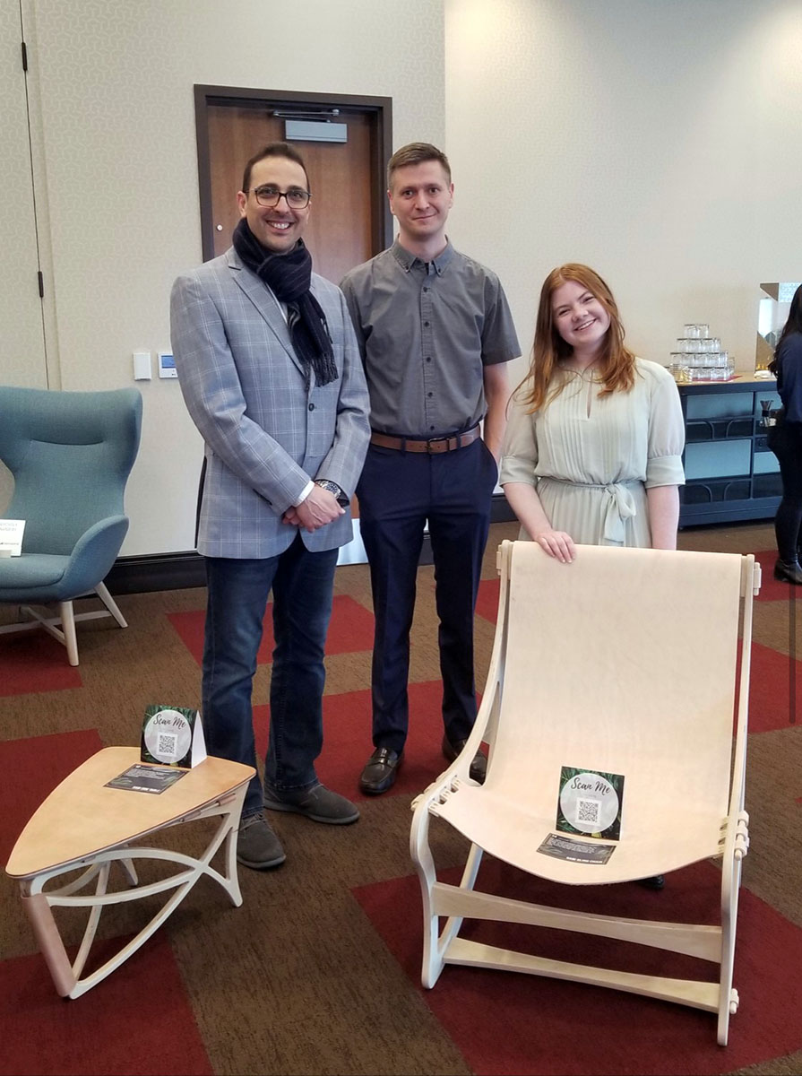 Two men and one woman posing next to table and chair.