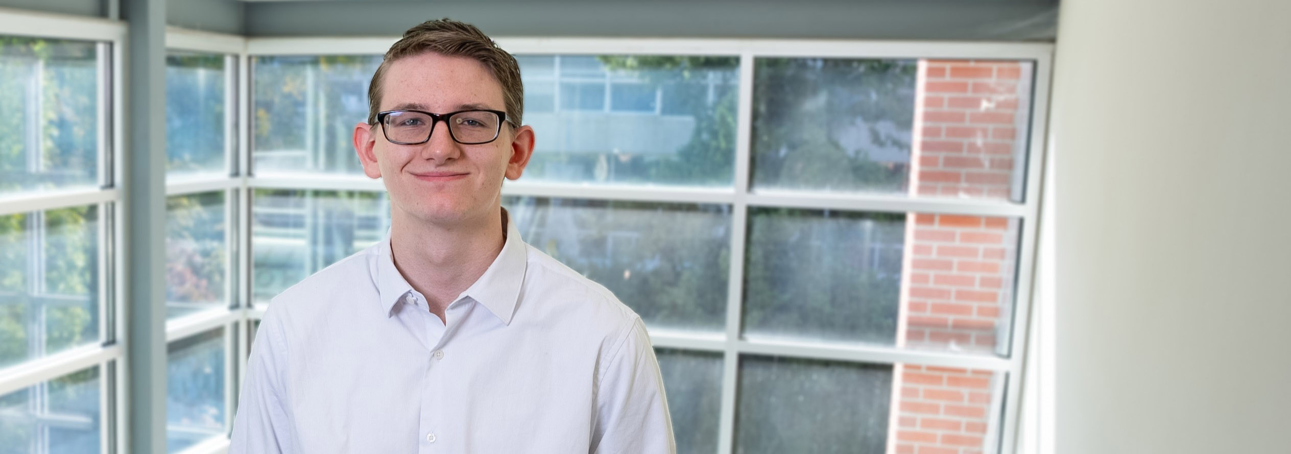 Young man with glasses stands in front of window.