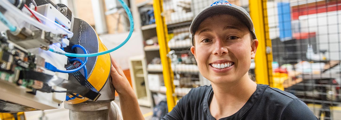 Smiling U of I student in NASA ball cap rests her hand on the arm of a large robot in a robotics lab.